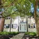 A two-story house with a white facade, black shutters, and a white front door, surrounded by lush green trees and a well-maintained garden.