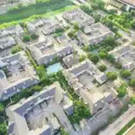 Aerial view of a residential neighborhood with multiple identical apartment buildings, arranged around parking lots and a central swimming pool, surrounded by green trees and lawns.