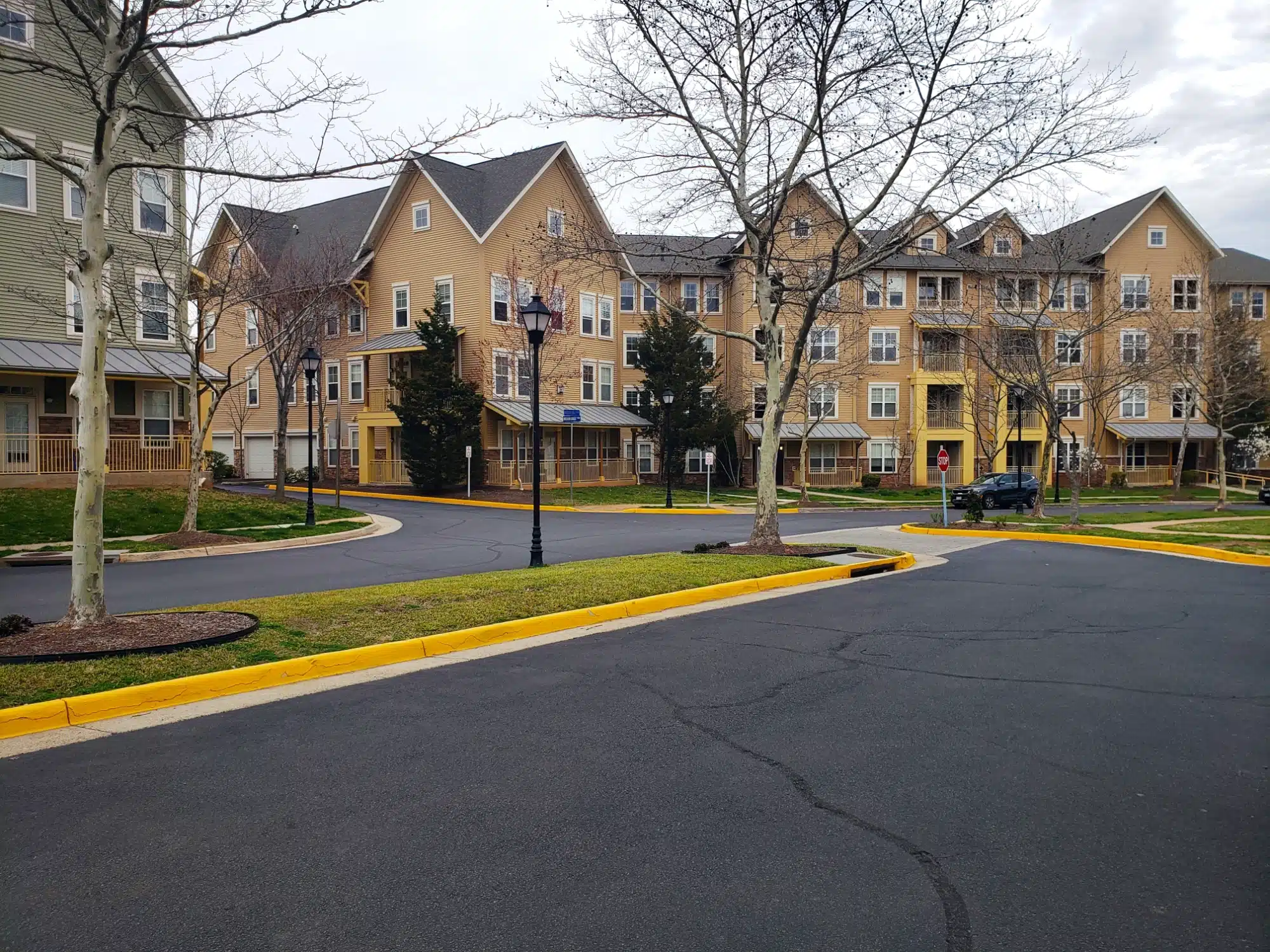 A complex of multi-story residential buildings with beige siding and white trim, surrounded by trees and a neatly paved road with yellow curbs. The setting appears calm and well-maintained, with streetlights and a parked car visible.