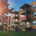 A brick apartment building with multiple balconies, seen in bright sunlight with a tree casting shadows on the grass.