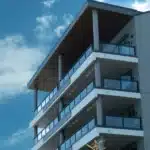 A modern apartment building with multiple glass-railed balconies and a flat roof, set against a clear blue sky with scattered clouds.