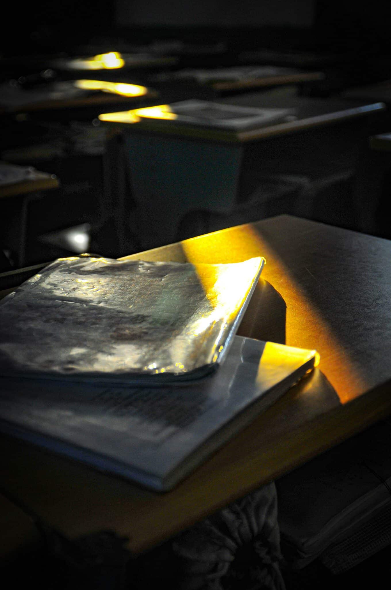 A classroom desk with a closed notebook illuminated by a beam of sunlight, with other desks visible in the shadowy background.