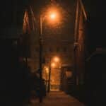A dark, snowy alleyway illuminated by a warm streetlight, with snowflakes visible against the night sky and buildings lining the sides.
