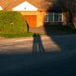 Shadows of two people on a street in front of a house, with a large bush and warm sunlight casting long shadows.