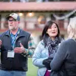 A group of four people are engaged in conversation outdoors. One man wearing a baseball cap and glasses gestures while speaking. A woman with long hair and a scarf listens attentively. They are outside a building with tile roofing, and the atmosphere appears informal and social.
