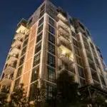 A multi-story urban apartment building with brick and glass facade, featuring several balconies, illuminated at night under a clear, starry sky.