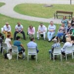 A group of people sitting in a circle on white chairs outdoors on a grassy area, engaged in a discussion.