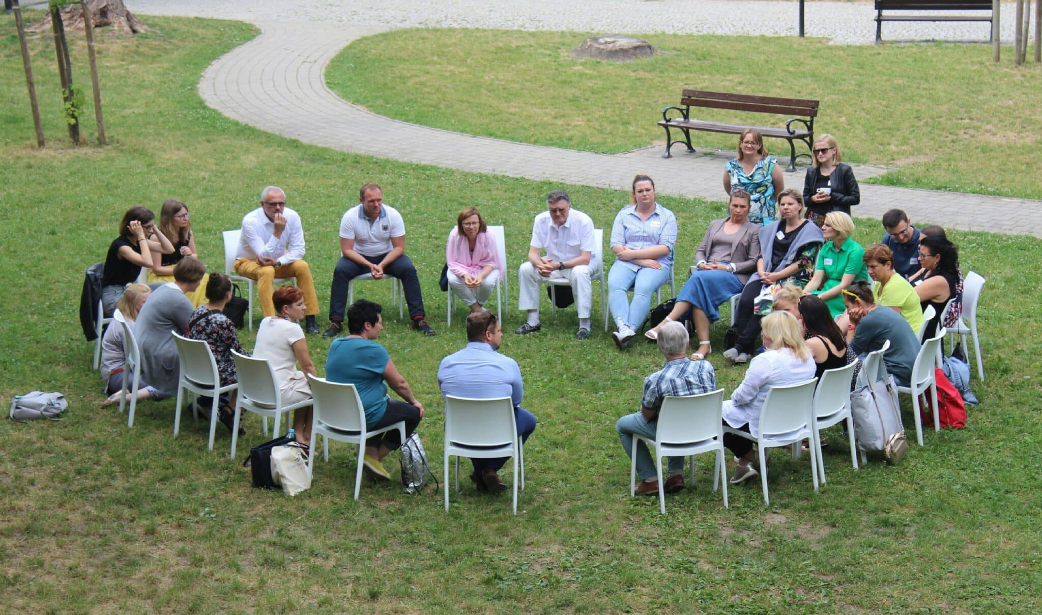 A group of people sitting in a circle on white chairs outdoors on a grassy area, engaged in a discussion.