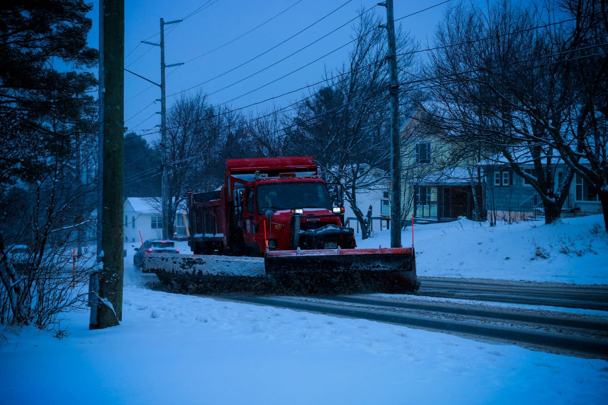 A red snow plow clearing snow from a residential street, with a car following behind. The scene is set in a snowy neighborhood during the evening, with trees and houses in the background.