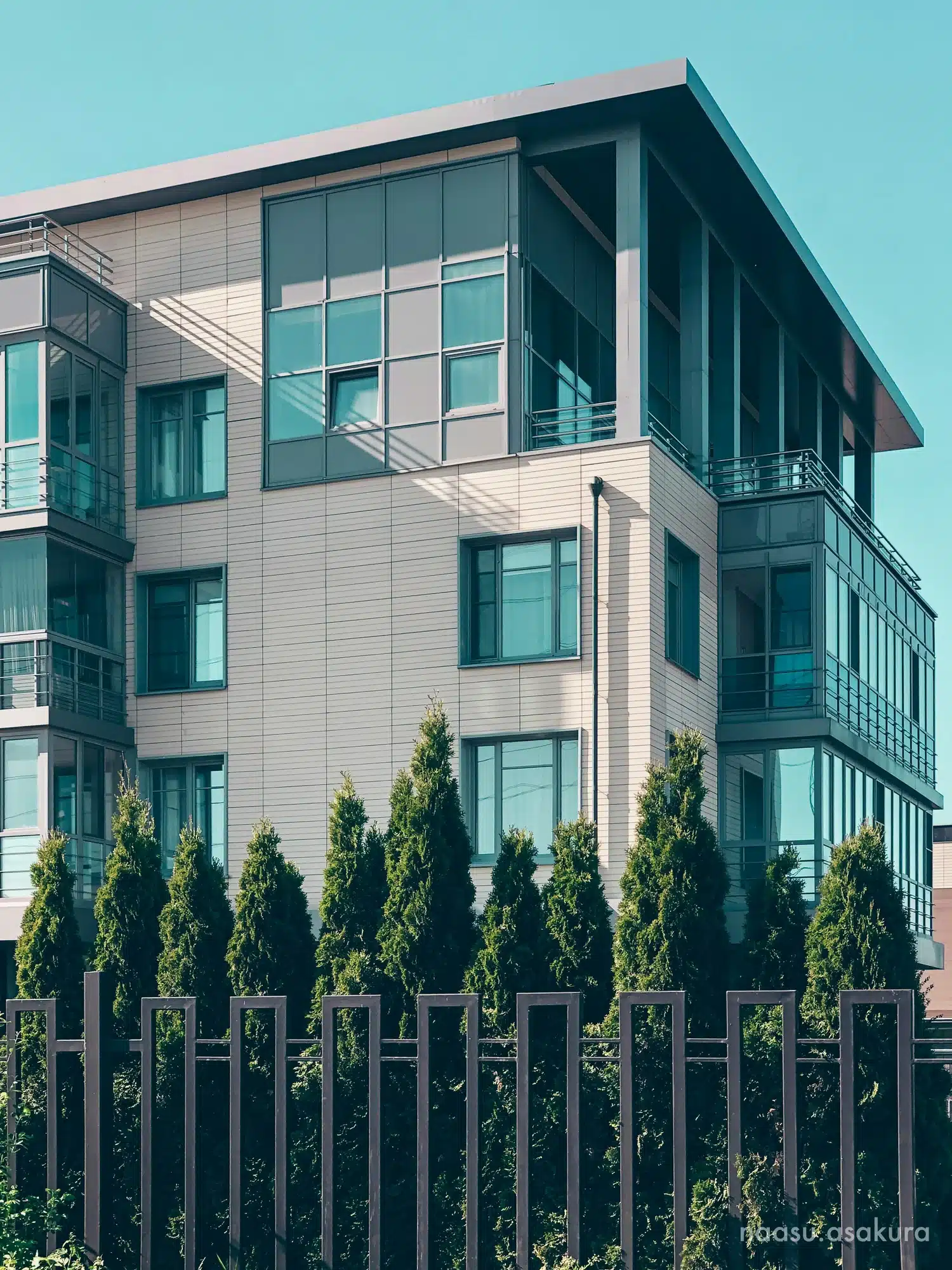 A modern apartment building with a sleek design and large windows, surrounded by tall evergreen shrubs and a metal fence, under a clear blue sky.